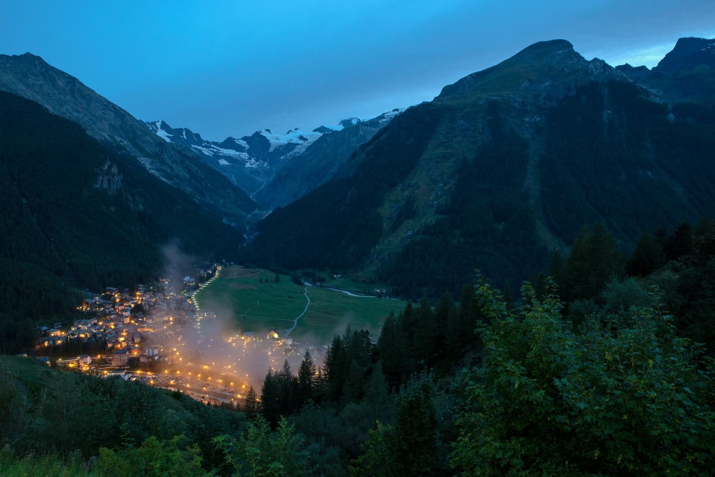 A lit village on the valley floor at dusk beneath dark mountain walls